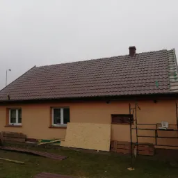 Dom jednorodzinny z częściowo wymienionym, brązowym dachem z blachodachówki; dwóch robotników w odblaskowych kamizelkach przygotowuje kolejne arkusze na trawniku, widoczne rusztowanie i płyta OSB