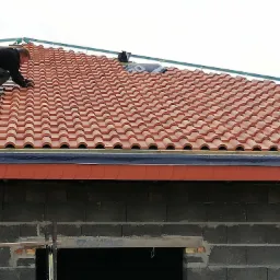 Dekarz układa czerwoną dachówkę ceramiczną na konstrukcji dachu nowego budynku z szarego bloczka, widoczna część rusztowania i niebieski pas pod dachówką.