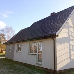 Dom jednorodzinny z grafitowym dachem z dachówki ceramicznej, brązowymi rynnami i jasną elewacją. Widoczne okna z białymi ramami i fragment trawnika z resztkami śniegu. Na dachu komin z cegły.