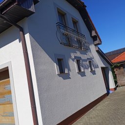 ZAKŁAD OGÓLNOBUDOWLANY JACEK WITEMBERG - Boczny widok domu z balkonem i brązową rynną, świeżo po renowacji elewacji. Widoczne trzy okna i fragment dachu z czerwoną dachówką.