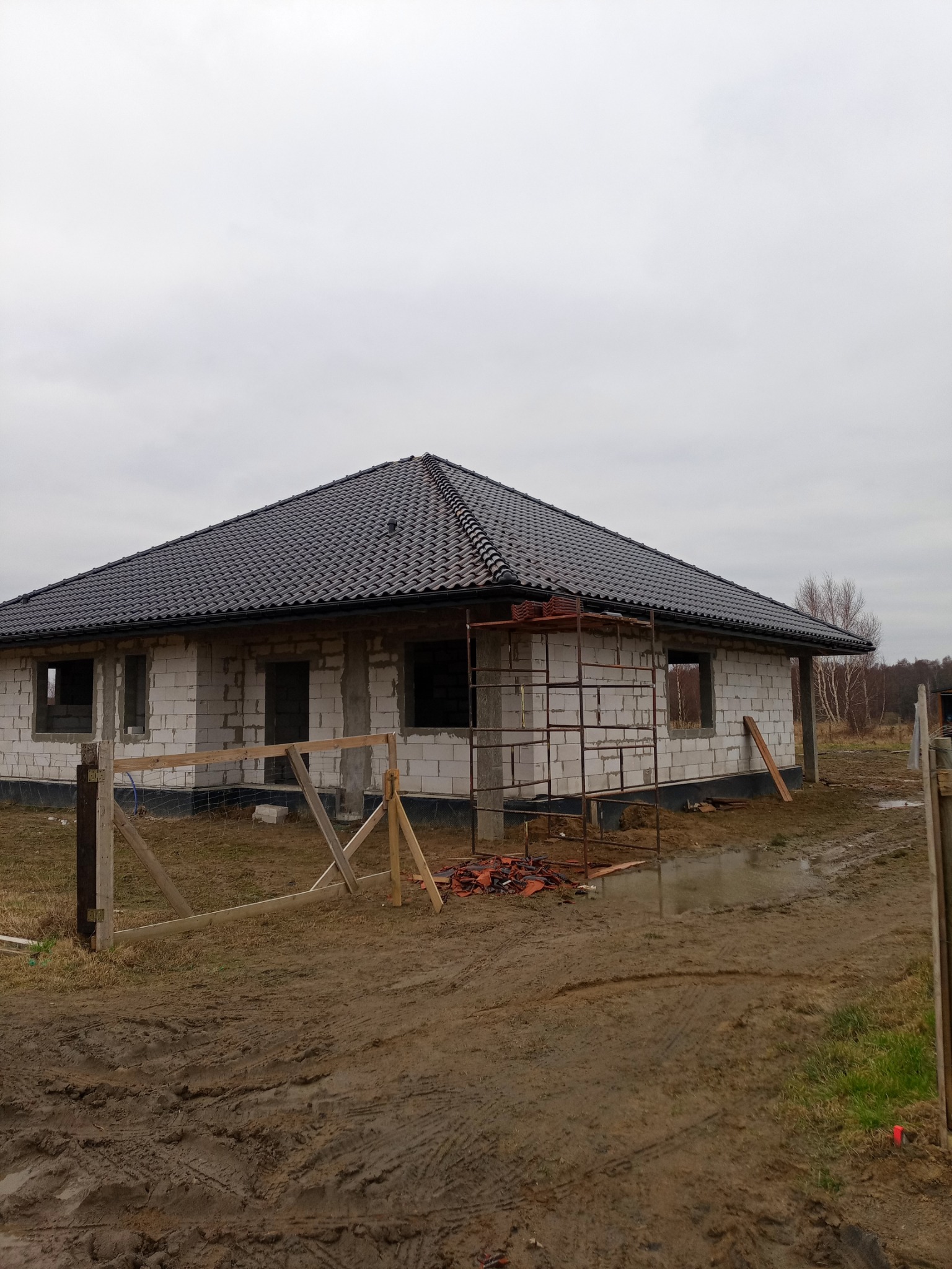 Dom w trakcie budowy z czarną dachówką, widoczne rusztowanie i porozrzucane dachówki, błotniste podłoże.
