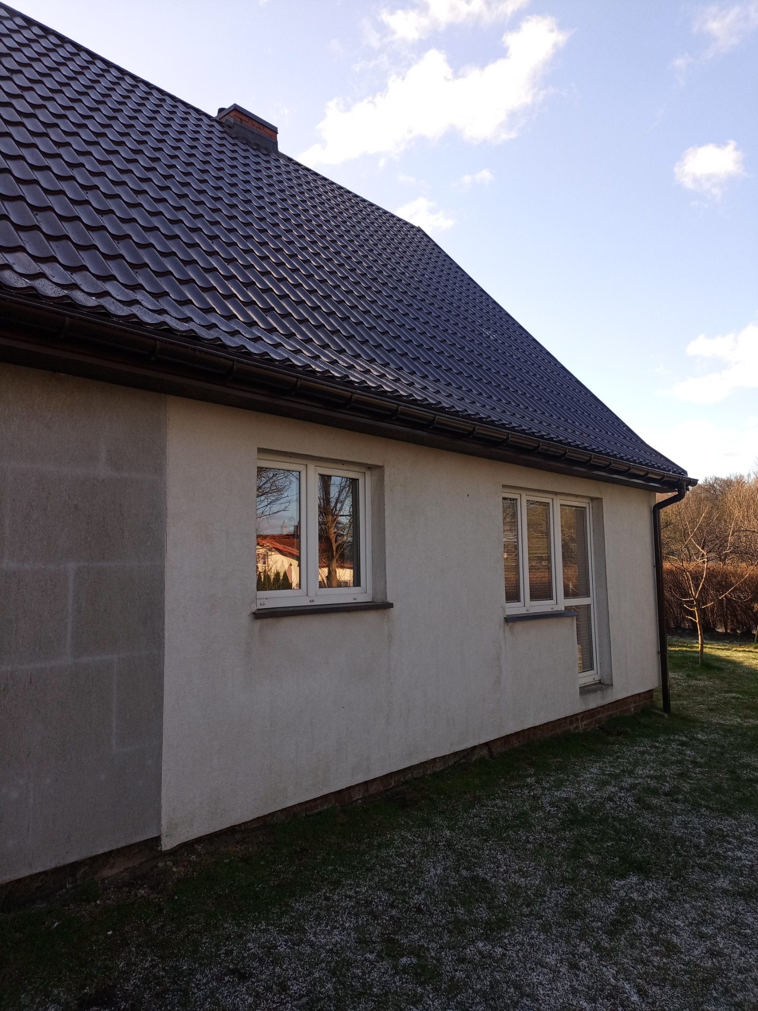 Dom jednorodzinny z ciemną, błyszczącą dachówką ceramiczną, widoczne rynny i okna z odbiciem sąsiedniego budynku. Ściany budynku otynkowane na jasno, z fragmentem ściany z pustaków. Na trawniku...