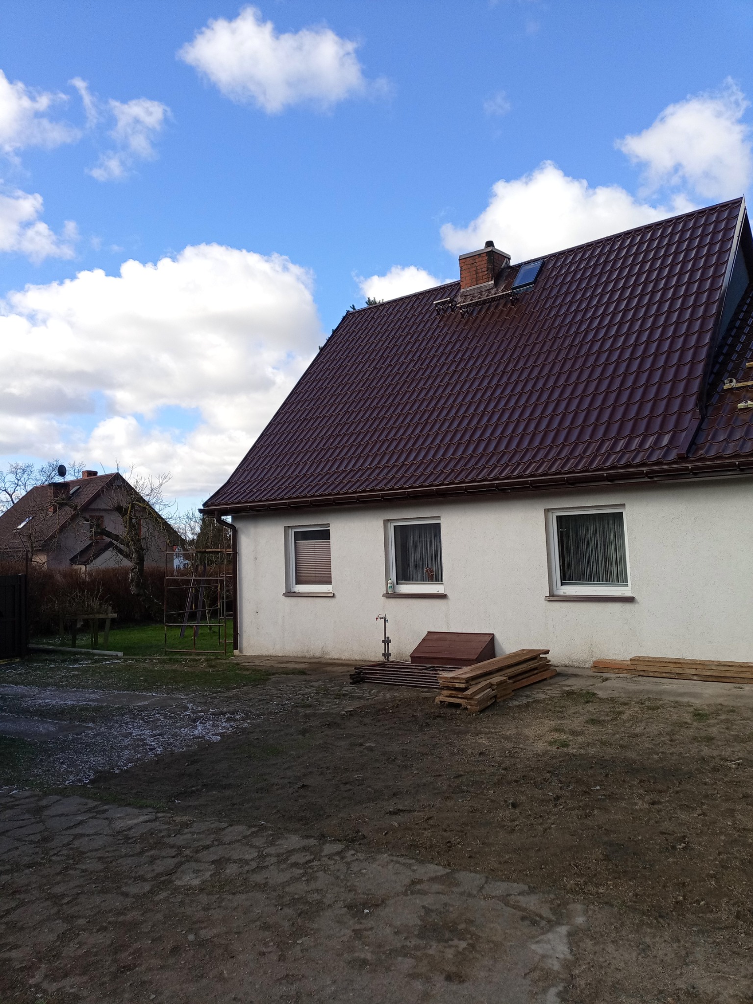 Dom jednorodzinny z nowym, błyszczącym, brązowym dachem z dachówki ceramicznej. Na dachu widoczny komin z cegły oraz okno dachowe. Przed budynkiem leżą deski i skrzynia.