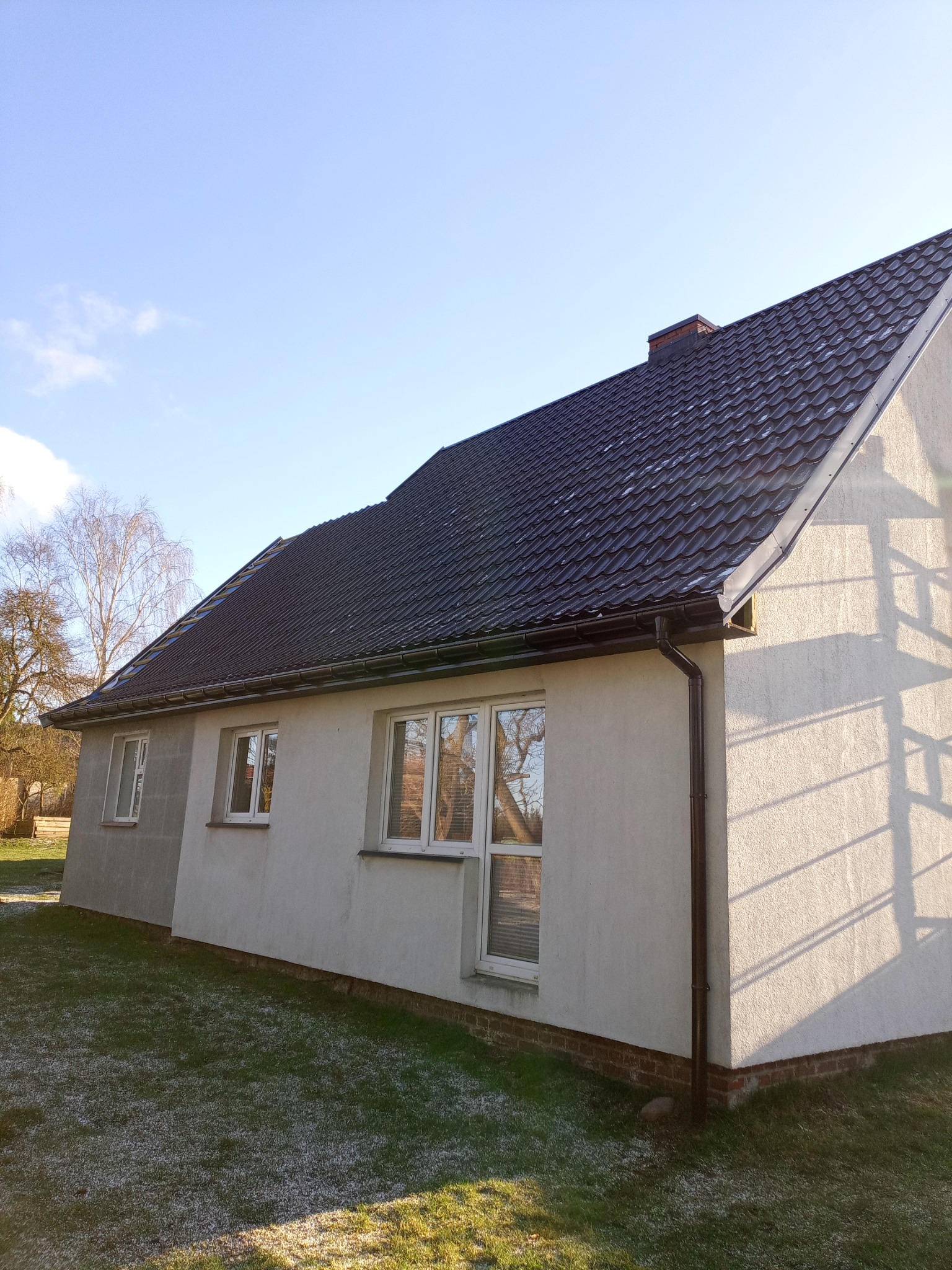 Dom jednorodzinny z grafitowym dachem z dachówki ceramicznej, brązowymi rynnami i jasną elewacją. Widoczne okna z białymi ramami i fragment trawnika z resztkami śniegu. Na dachu komin z cegły.