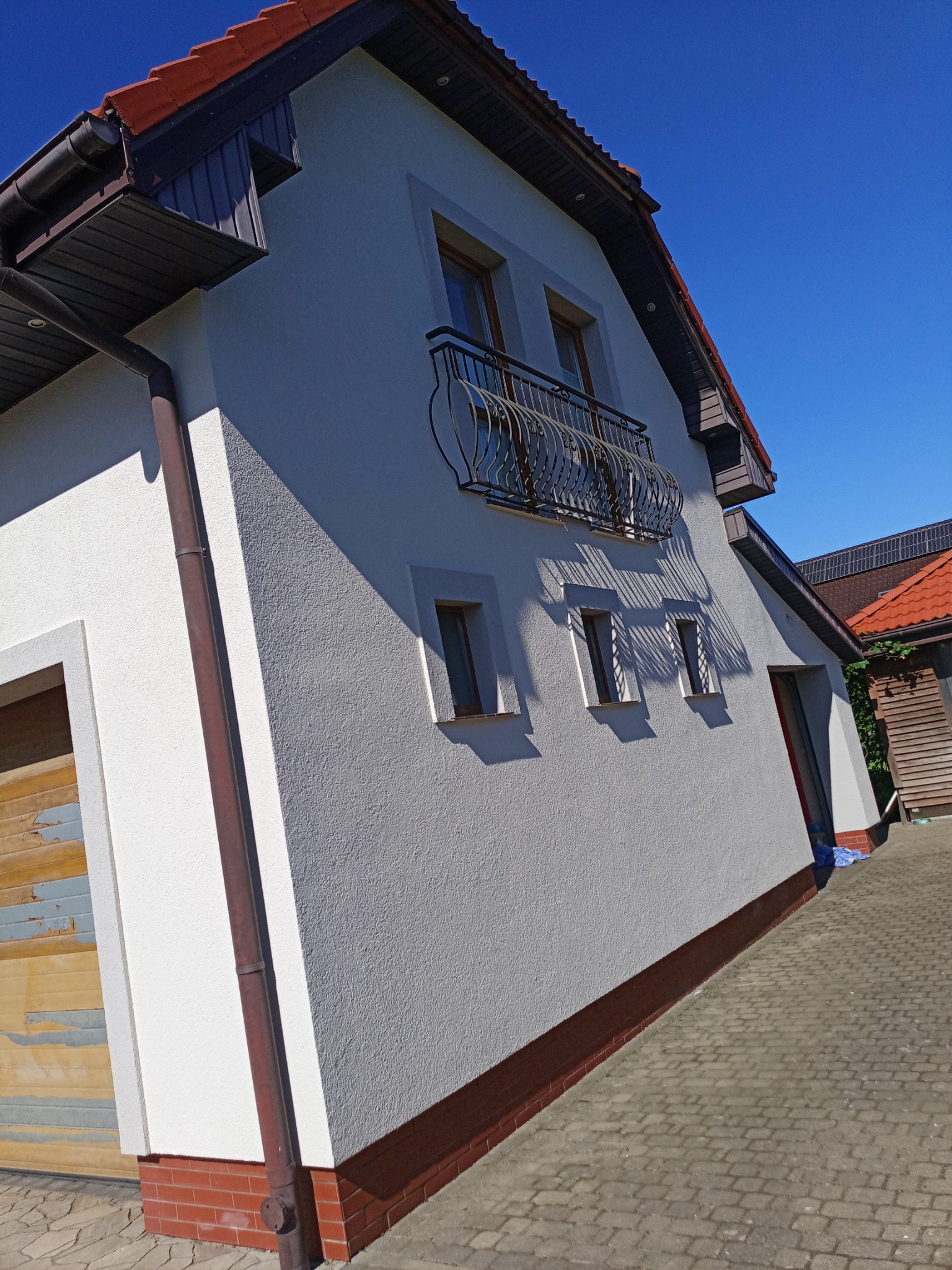 Boczny widok domu z balkonem i brązową rynną, świeżo po renowacji elewacji. Widoczne trzy okna i fragment dachu z czerwoną dachówką.