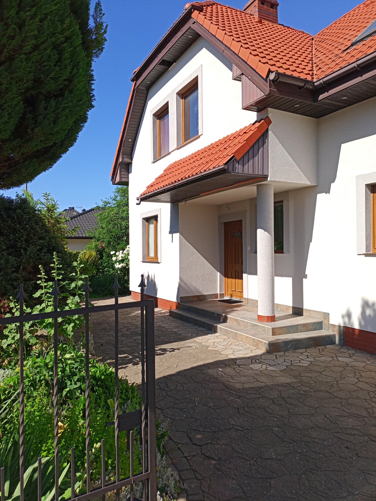 Jasny dom jednorodzinny z czerwonym dachem, widok zza metalowego ogrodzenia w słoneczny dzień. Elewacja w odcieniach bieli, brązowe okna i drzwi wejściowe.