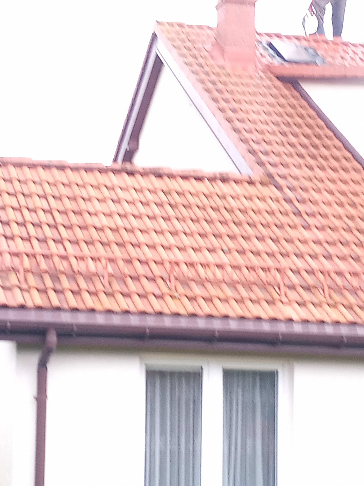 Częściowy widok domu z czerwoną dachówką, brązowymi rynnami i białymi ścianami. Na dachu widoczna osoba podczas pracy. Ujęcie z dołu.
