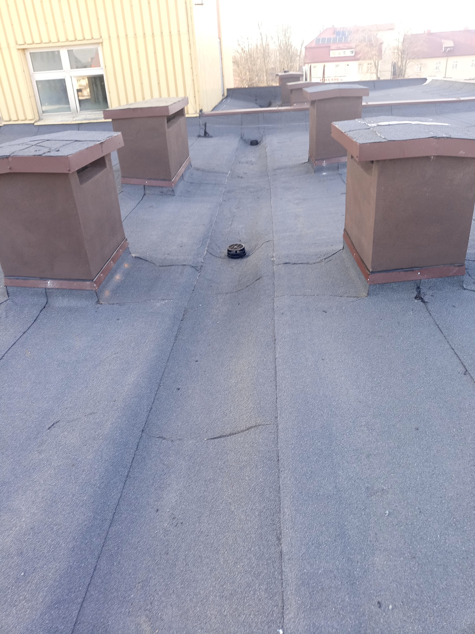 Płaski dach pokryty papą z brązowymi kominami wentylacyjnymi. Widoczne łączenia papy i elementy wentylacyjne. Budynek w tle.