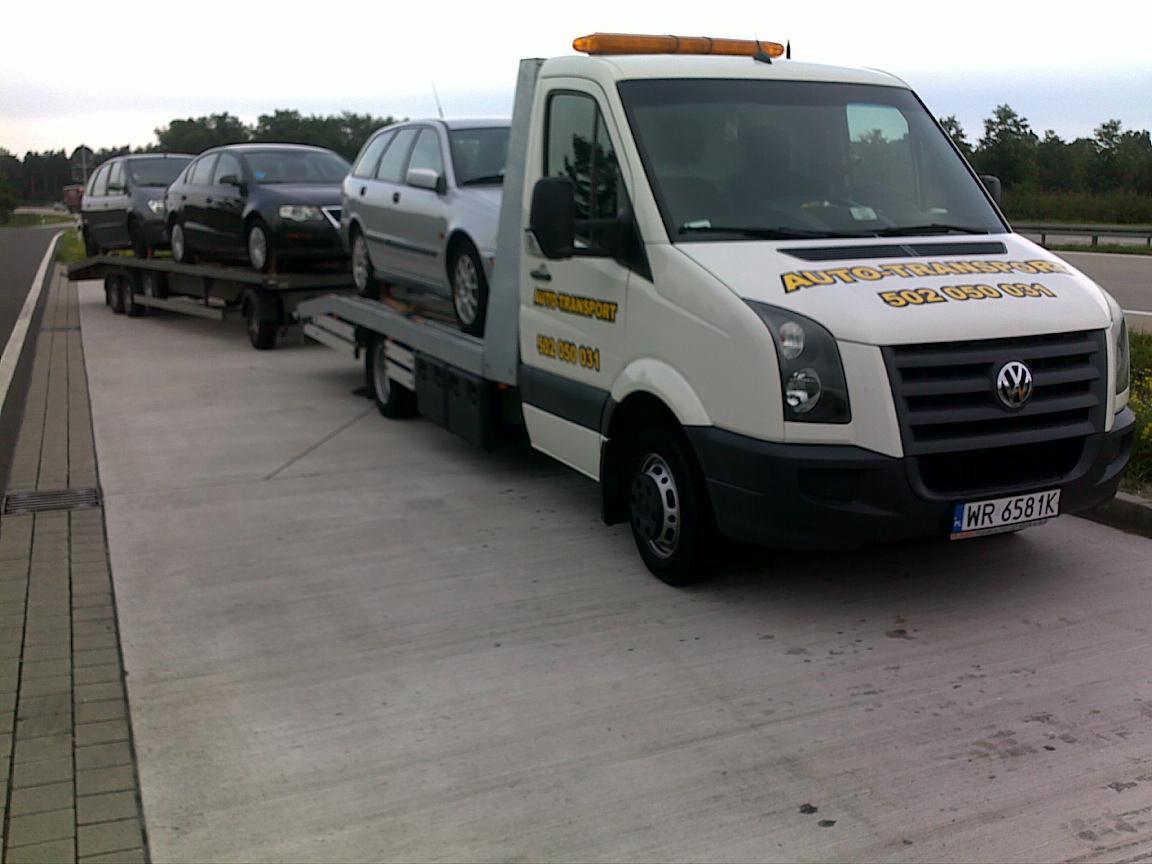 Biała laweta Volkswagen z trzema samochodami na platformie, zaparkowana na betonowym placu przy drodze, widoczne numery telefonu na kabinie.