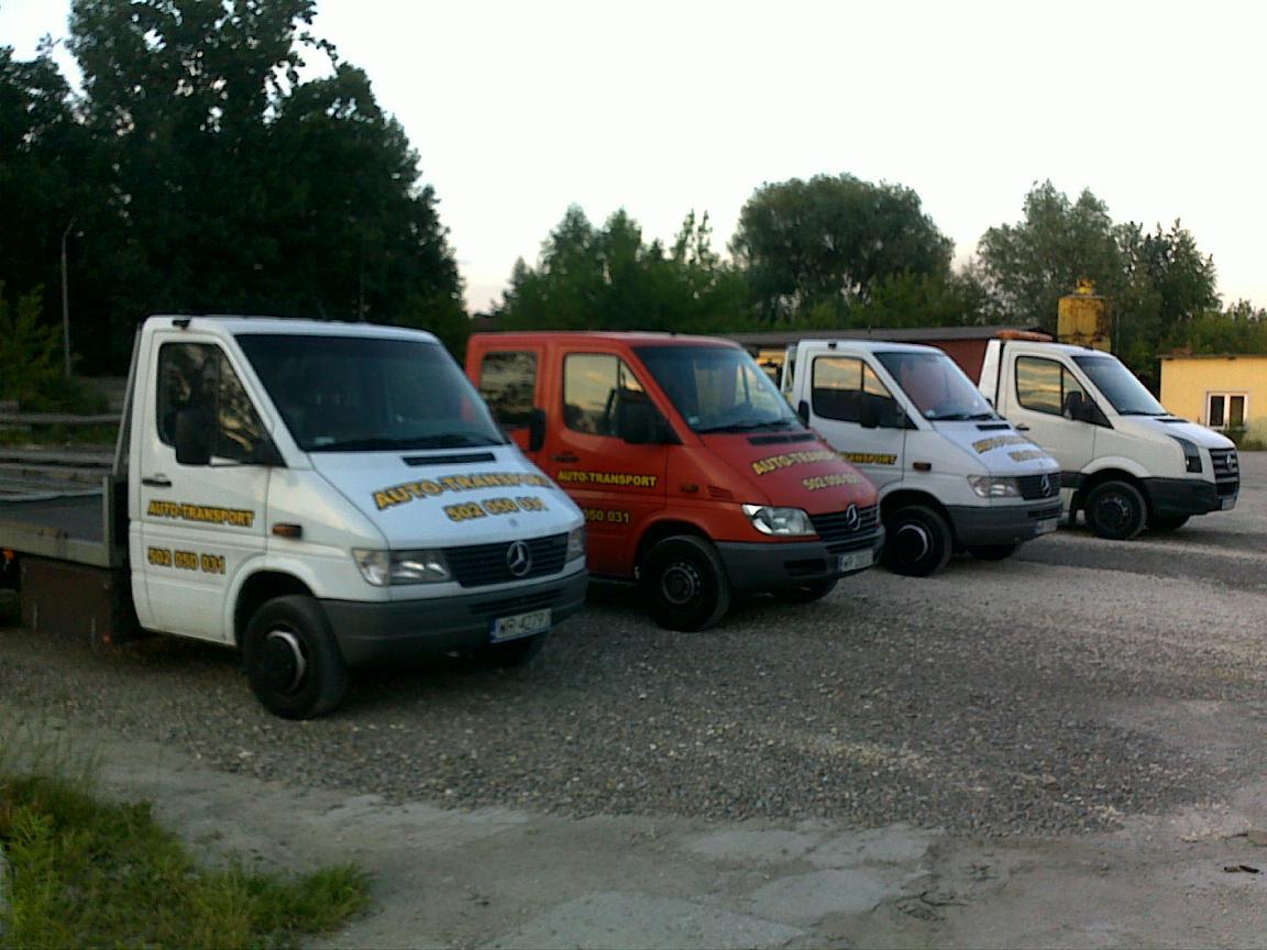 Flota lawet Mercedes-Benz Sprinter w kolorach białym i czerwonym, zaparkowana na żwirowym placu z widocznym napisem 'AUTO-TRANSPORT' i numerem telefonu na karoserii, w tle zieleń drzew i budynek.