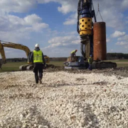 Prace ziemne z użyciem wiertnicy do wzmacniania gruntu pod budowę, widoczni pracownicy w kamizelkach odblaskowych i kaskach, na pierwszym planie wysypany tłuczeń.