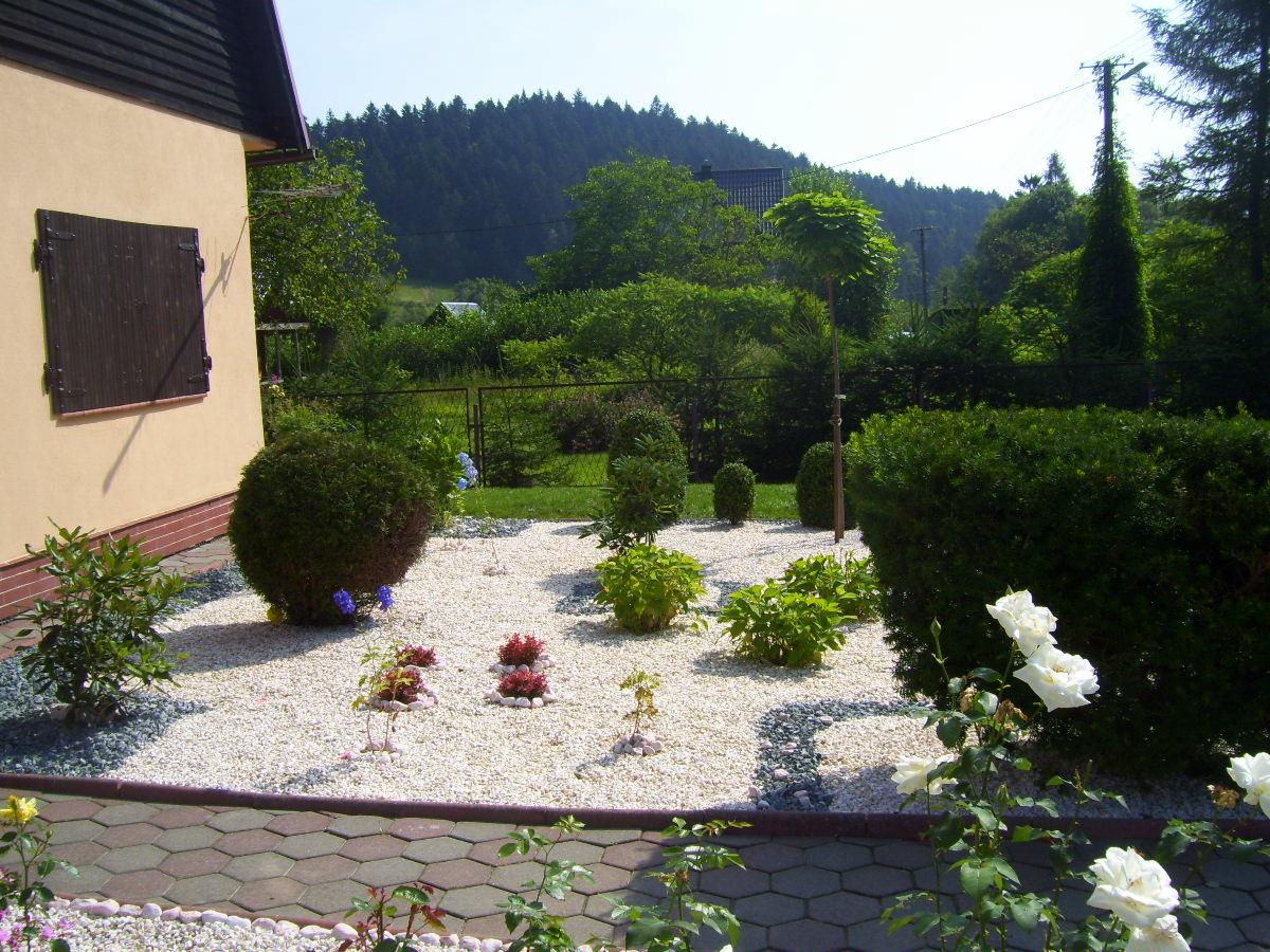 Ogród przydomowy z białym żwirem, ozdobnymi krzewami i kwiatami, widok z poziomu chodnika, w tle zalesione wzgórze.