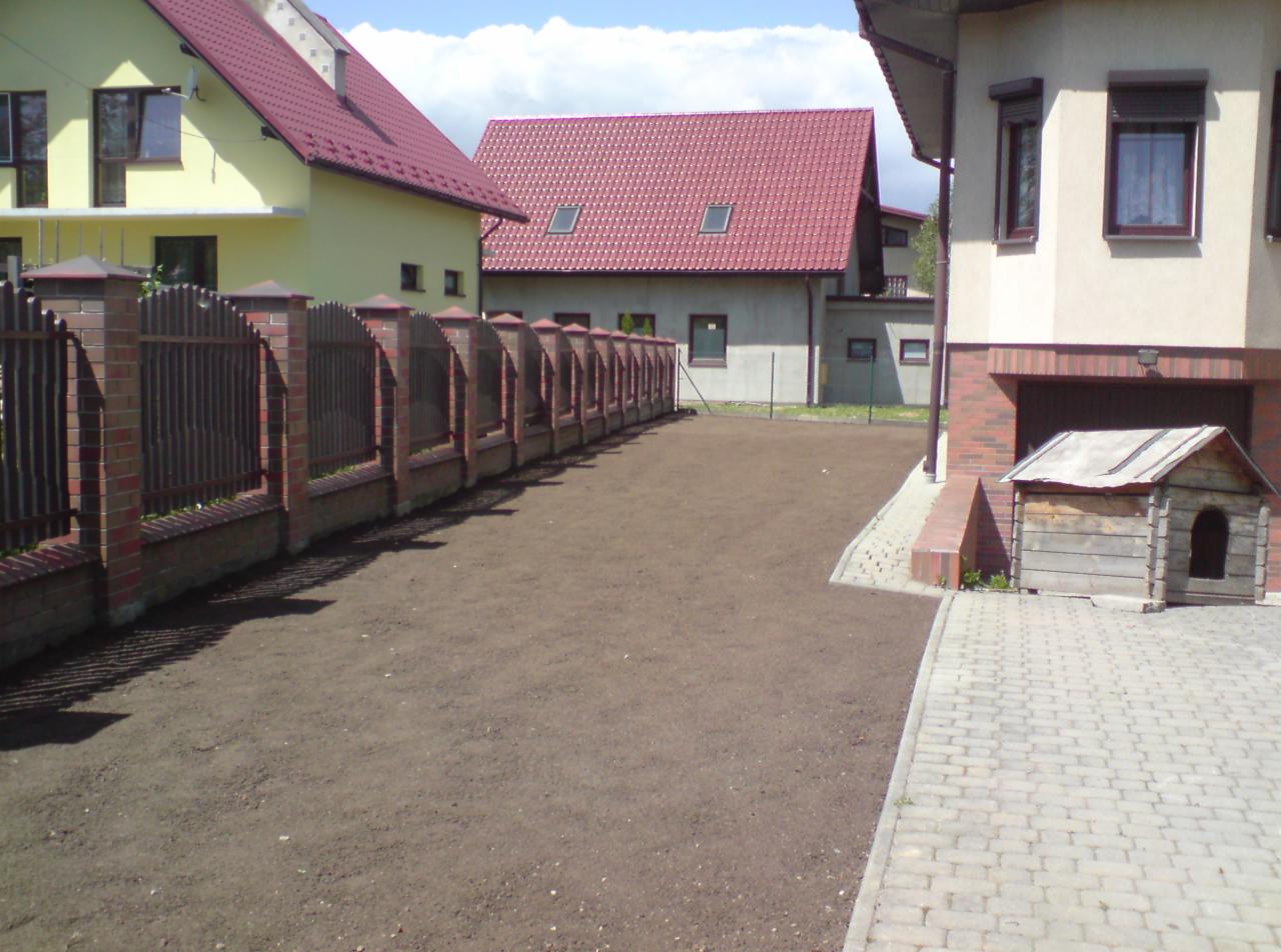 Przygotowany teren pod trawnik, z widoczną ziemią, obok kostki brukowej i budy dla psa, w tle domy z czerwonymi dachami i ceglany płot.