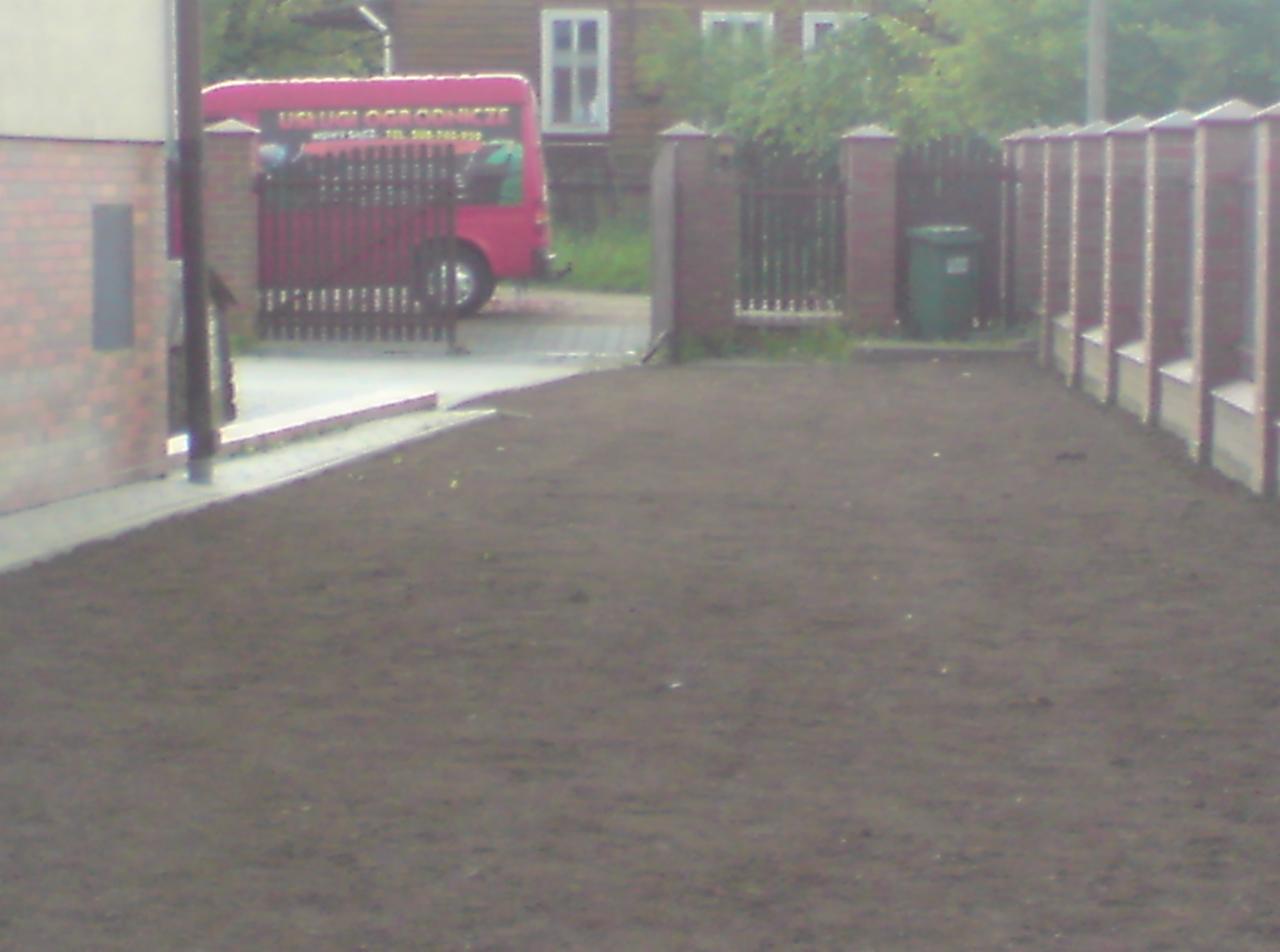 Przygotowany teren pod trawnik z widoczną ziemią, ogrodzeniem i samochodem firmy ogrodniczej w tle.