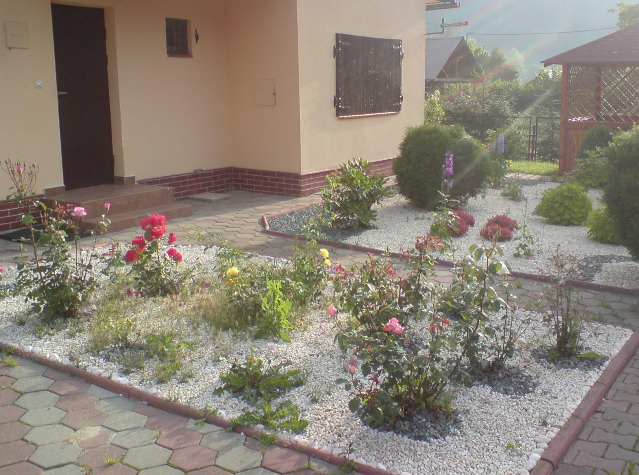 Ogród przed domem z rabatami wysypanymi białym żwirem, różnorodność róż w kolorach różowym, czerwonym i żółtym, obrzeża rabat z kostki, widoczny fragment domu z brązowymi okiennicami i fragment...