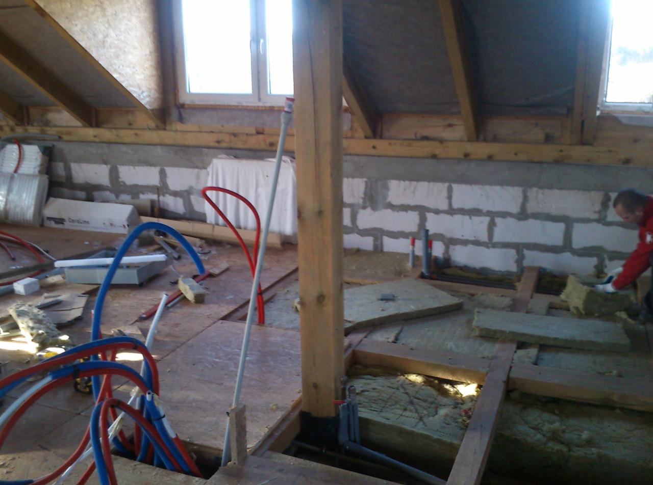 Montaż instalacji grzewczej podłogowej na poddaszu: rury w kolorach czerwonym i niebieskim, izolacja z wełny mineralnej, widoczny pracownik w czerwonej bluzie.