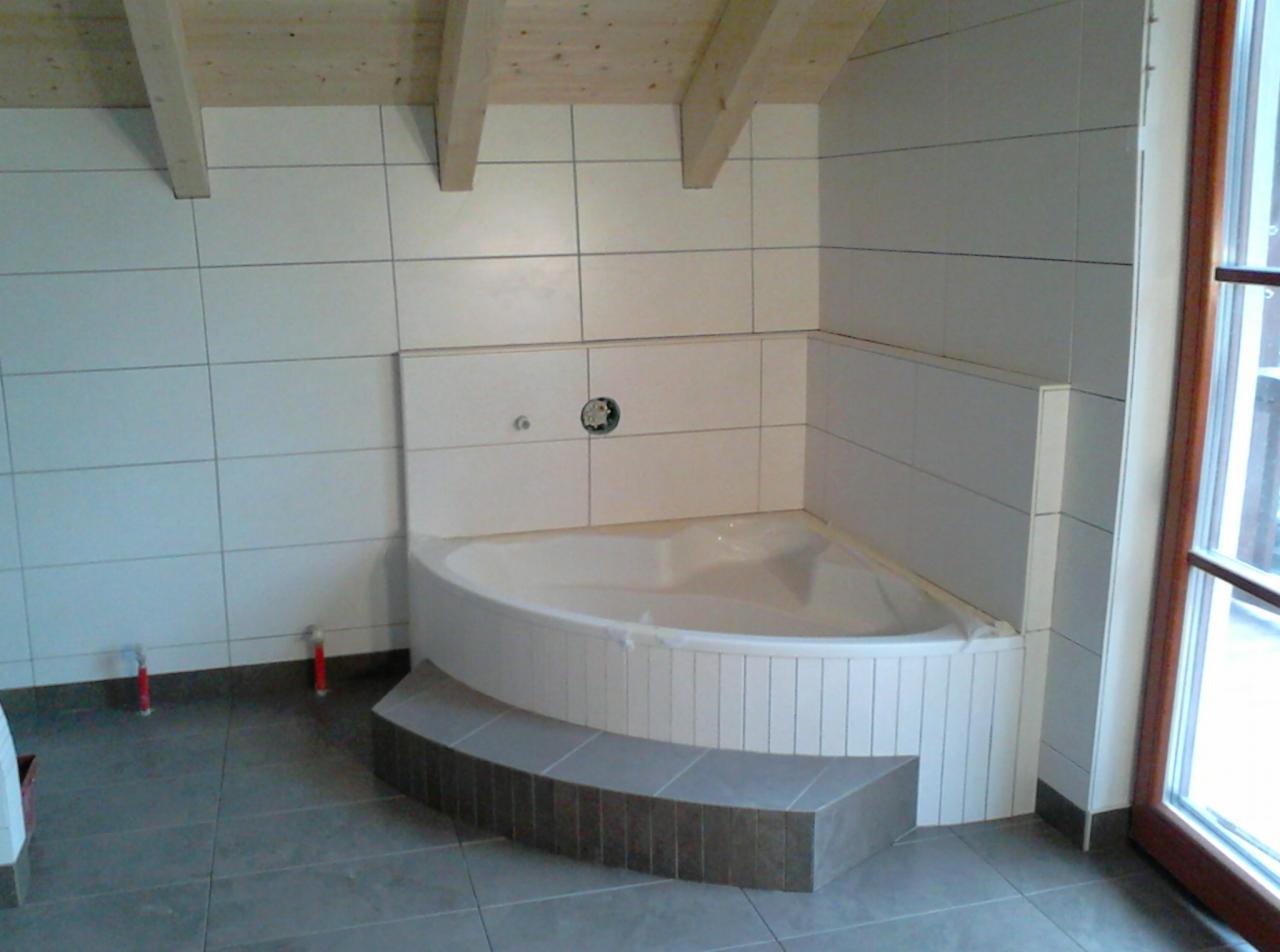 Wykończona łazienka w poddaszu: narożna wanna z obudową z białych płytek, szara podłoga i ściany wyłożone białymi płytkami. Widoczne drewniane belki stropowe.