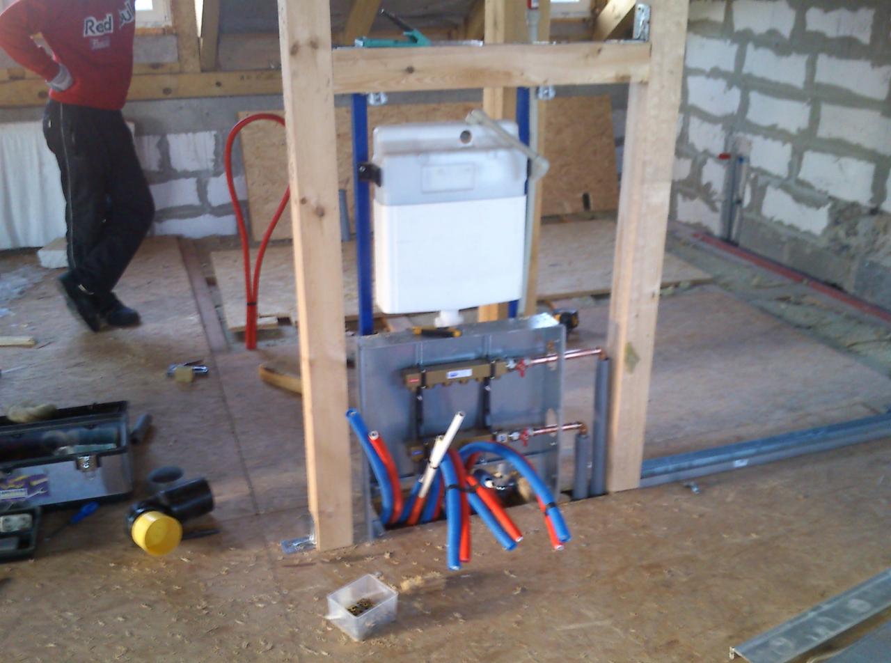 Stelaż podtynkowy WC z instalacją wodną w surowym wnętrzu. Widoczne rury PEX w kolorach niebieskim i czerwonym. Częściowo wykończona ściana z białej cegły.