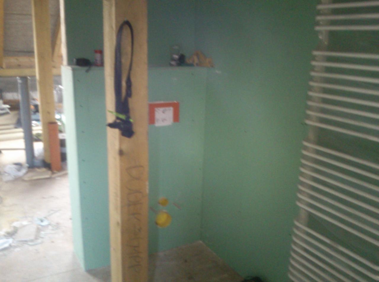 Wnętrze w trakcie budowy/remontu z zielonymi ścianami, widoczną instalacją i grzejnikiem drabinkowym. Elementy drewniane w konstrukcji. Ślady prac budowlanych na podłodze.