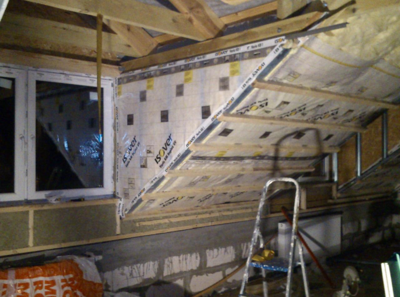Poddasze w trakcie izolacji, widoczna wełna mineralna, folia paroizolacyjna Isover, drewniane elementy konstrukcyjne i drabina. Okno po lewej stronie.