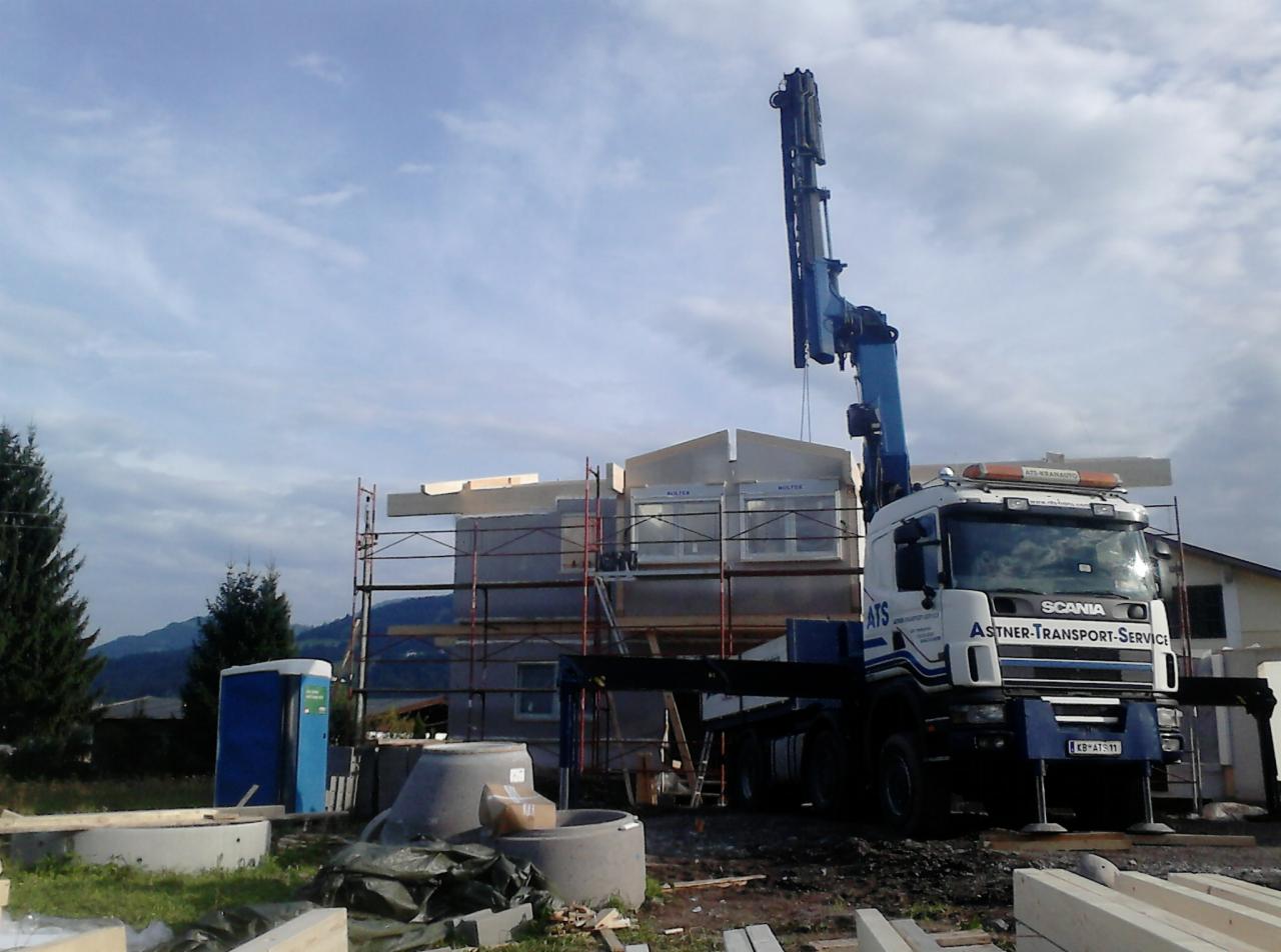 Montaż prefabrykowanego domu modułowego z użyciem dźwigu samochodowego. Na pierwszym planie betonowe kręgi szamba i przenośna toaleta.