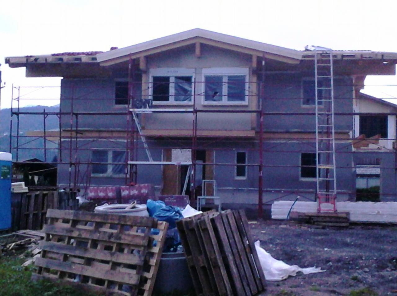 Budowa domu dwukondygnacyjnego w trakcie ocieplania, z rusztowaniami i paletami na pierwszym planie. W tle widoczne góry.