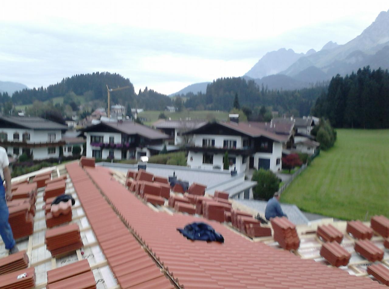 Układanie czerwonej dachówki na dachu z widokiem na góry i zabudowania, w tle las. Częściowo ułożony dach, przygotowane paczki dachówek. Praca na wysokości.