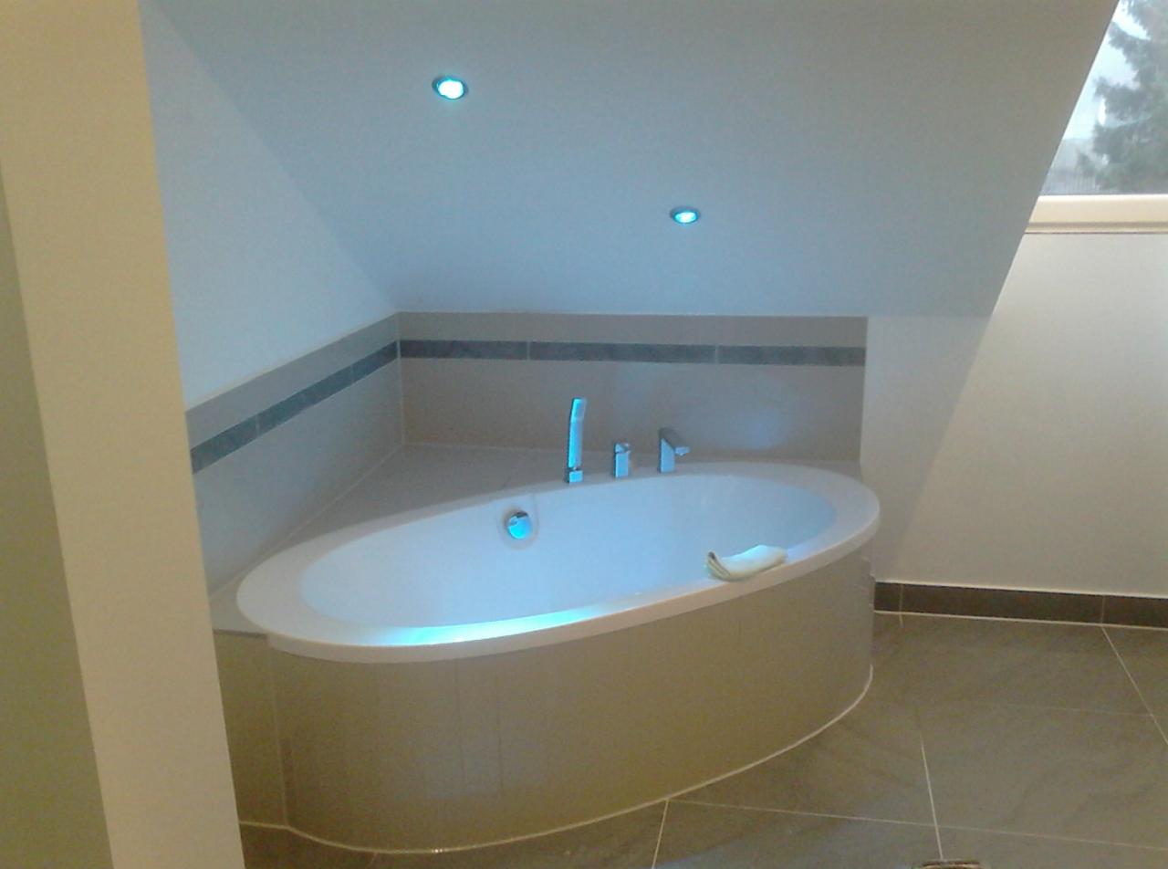 Nowoczesna, owalna wanna z hydromasażem w wykafelkowanej obudowie, umieszczona w łazience na poddaszu z oknem i oświetleniem punktowym w suficie.