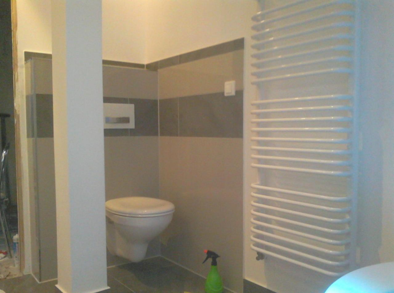 Nowoczesna łazienka z podwieszaną toaletą, grzejnikiem drabinkowym i szaro-beżowymi płytkami. Widoczna butelka ze środkiem czyszczącym na podłodze.