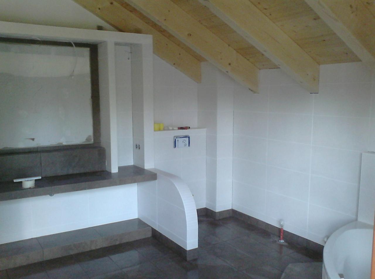 Nowoczesna łazienka na poddaszu z drewnianą więźbą dachową, wykończona białymi płytkami i ciemną podłogą. Widoczna zabudowa umywalki i fragment wanny.