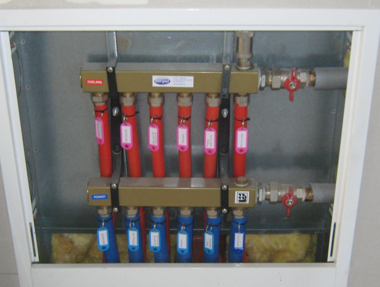 Rozdzielacz ogrzewania podłogowego w metalowej szafce z izolacją, rury czerwone i niebieskie z etykietami, zawory kulowe, instalacja wewnątrz budynku.