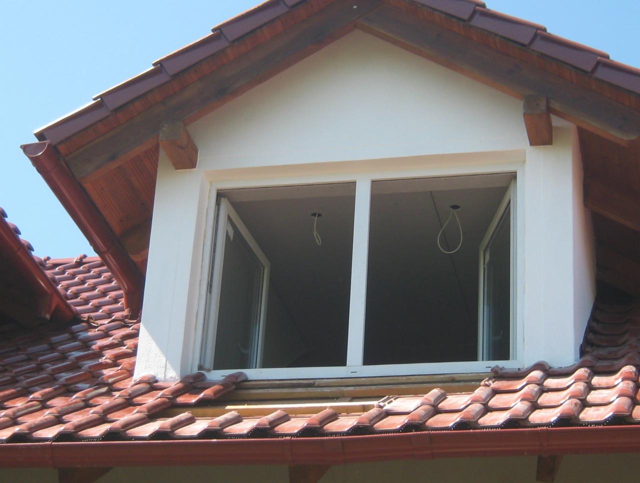 Montaż okna dachowego w Zielonej Górze. Widok z zewnątrz na białe okno PCV na poddaszu z czerwoną dachówką. Widoczne kable elektryczne.
