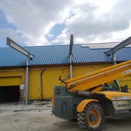 Montaż stalowych belek do niebieskiego dachu z blachy trapezowej, pracownik na drabinie przy żółtej ścianie budynku, w tle widoczny podnośnik teleskopowy.