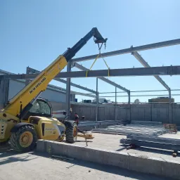 Żółty wózek widłowy New Holland podnosi stalową konstrukcję dachu na placu budowy, widoczny pracownik w tle.