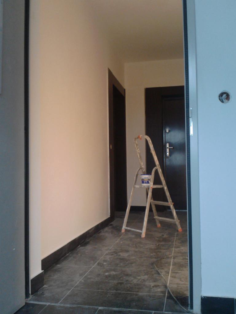 Świeżo pomalowany korytarz z widoczną drabiną i puszką farby, czarne płytki podłogowe i ciemne listwy przypodłogowe.