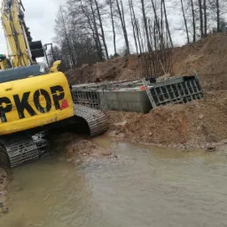 Żółta koparka PKOP obok fundamentu zbrojonego, w wykopie z wodą. Widoczne szalunki i stalowe pręty. Tło to drzewa i ziemia. Pochmurny dzień.