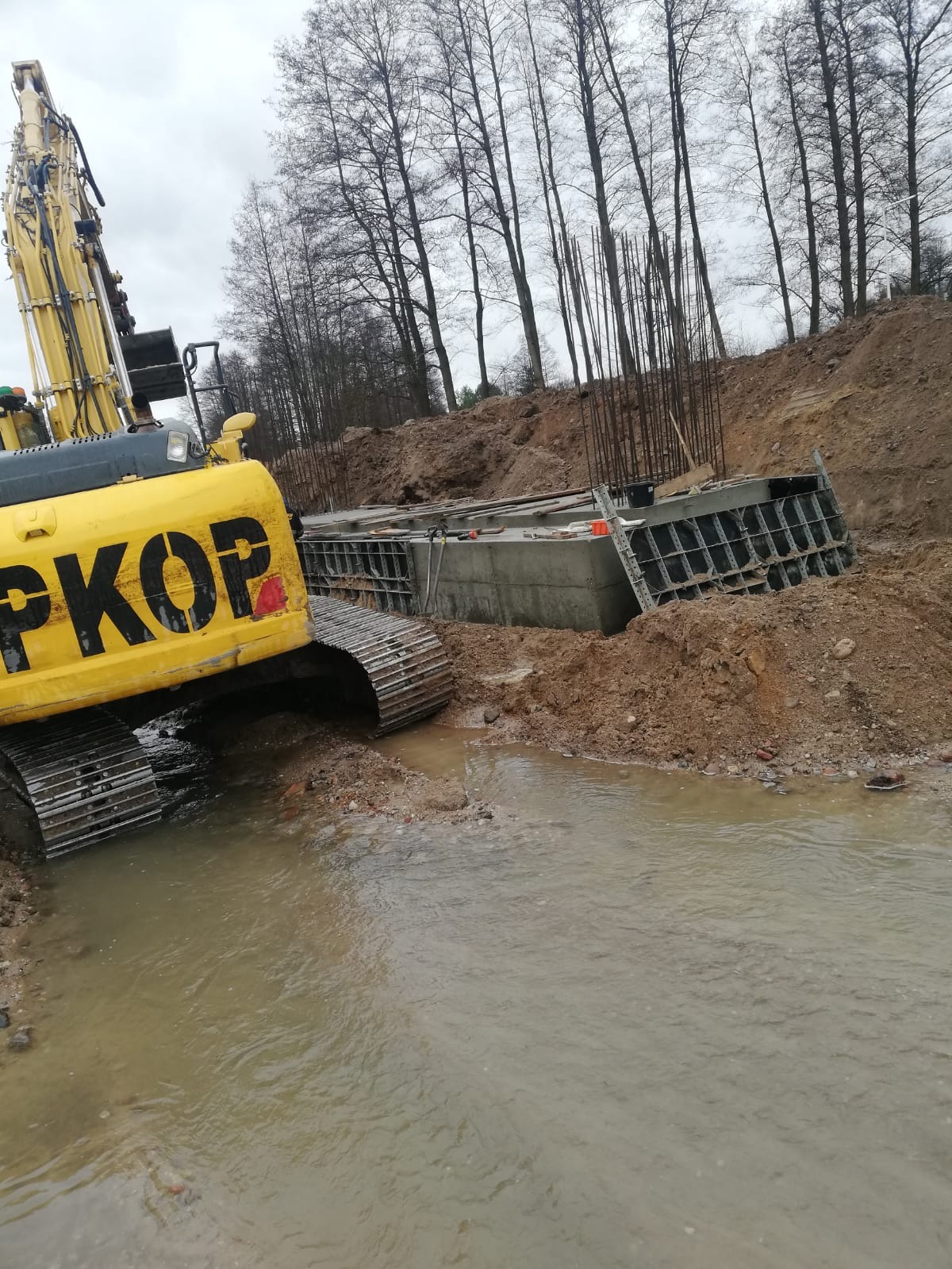 Żółta koparka PKOP obok fundamentu zbrojonego, w wykopie z wodą. Widoczne szalunki i stalowe pręty. Tło to drzewa i ziemia. Pochmurny dzień.