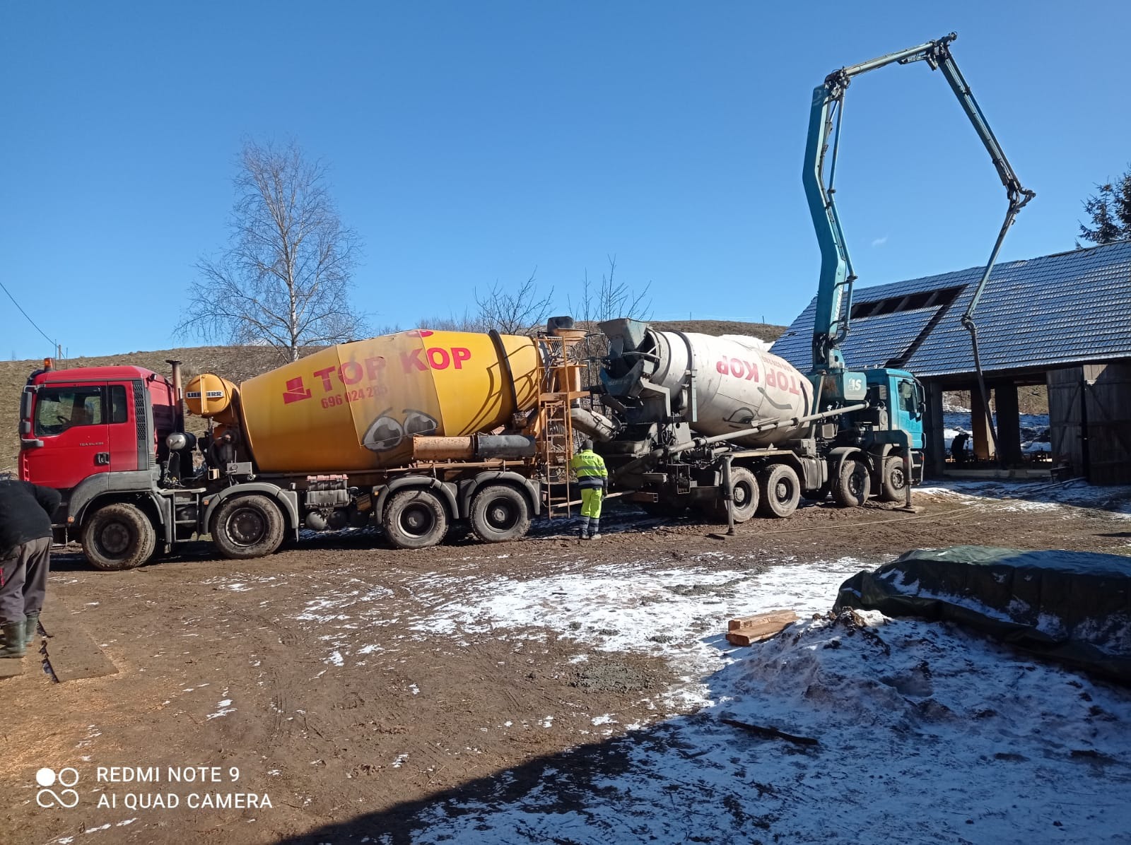 Dwa betonomieszarki 'TOP KOP' na placu budowy, z pracownikiem w odblaskowej kamizelce. W tle budynek z niebieskim dachem i resztki śniegu na ziemi.