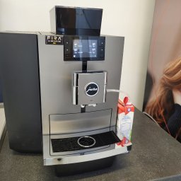 FIRA Robert Firkowski - Profesjonalny ekspres do kawy Jura X10 w biurowym otoczeniu, obok karton mleka i plakat z wizerunkiem kobiety.