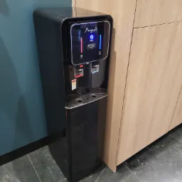 Czarny, wolnostojący dystrybutor wody z wyświetlaczem LED, umieszczony obok drewnianych szafek kuchennych i niebieskiej ściany.