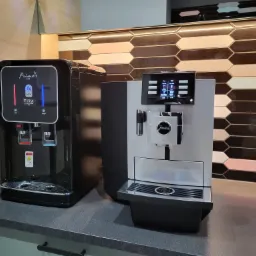 Czarny dystrybutor wody Prime FIRA z podświetleniem LED obok srebrnego ekspresu do kawy Jura XS na blacie z ciemnego kamienia, na tle ściany z dekoracyjnymi płytkami w kształcie rombów w odcieniach...