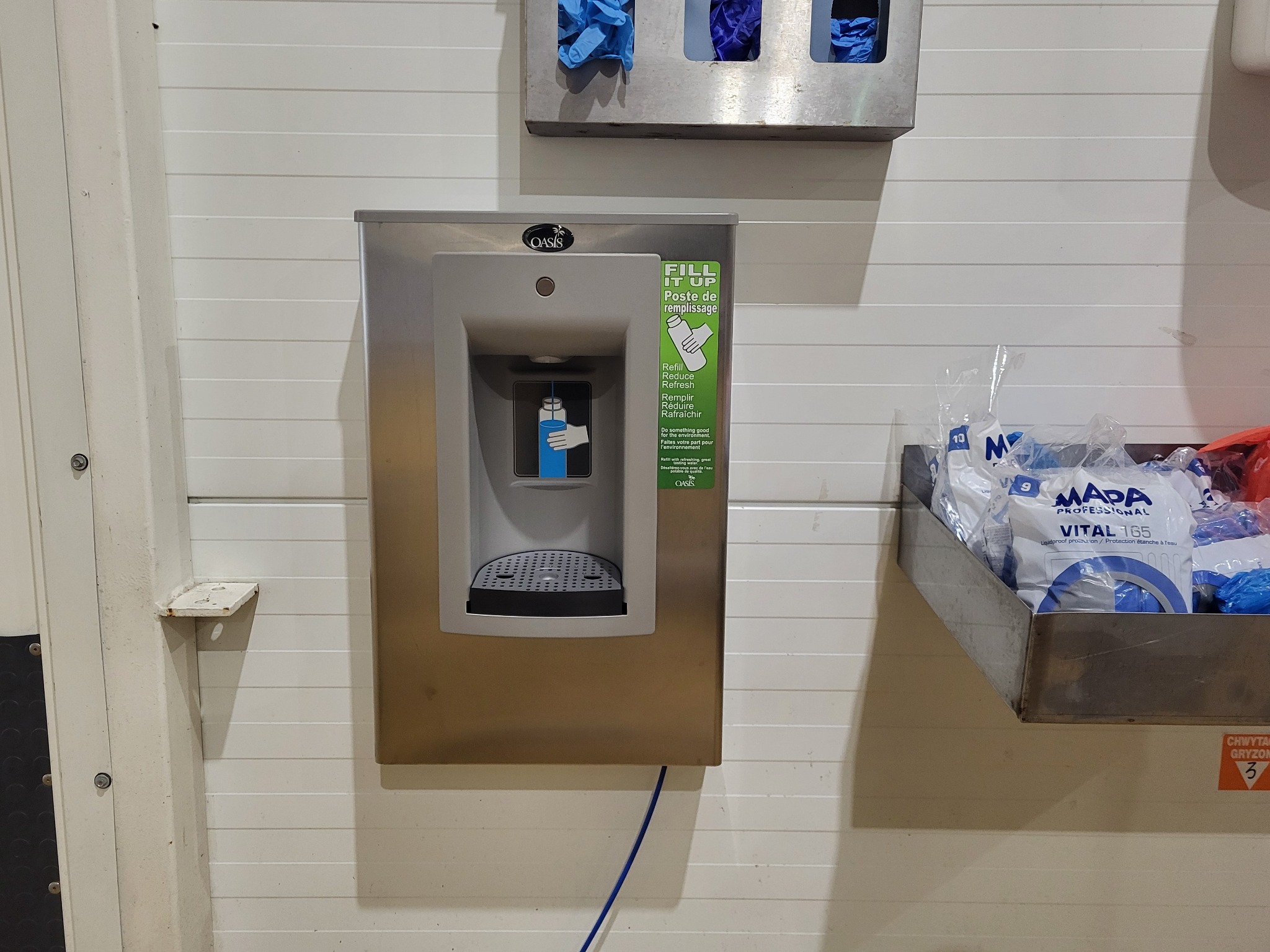 Srebrny dystrybutor wody Oasis na ścianie, z naklejką informującą o możliwości napełniania butelek i oszczędzaniu środowiska, obok pojemnik z rękawicami ochronnymi.