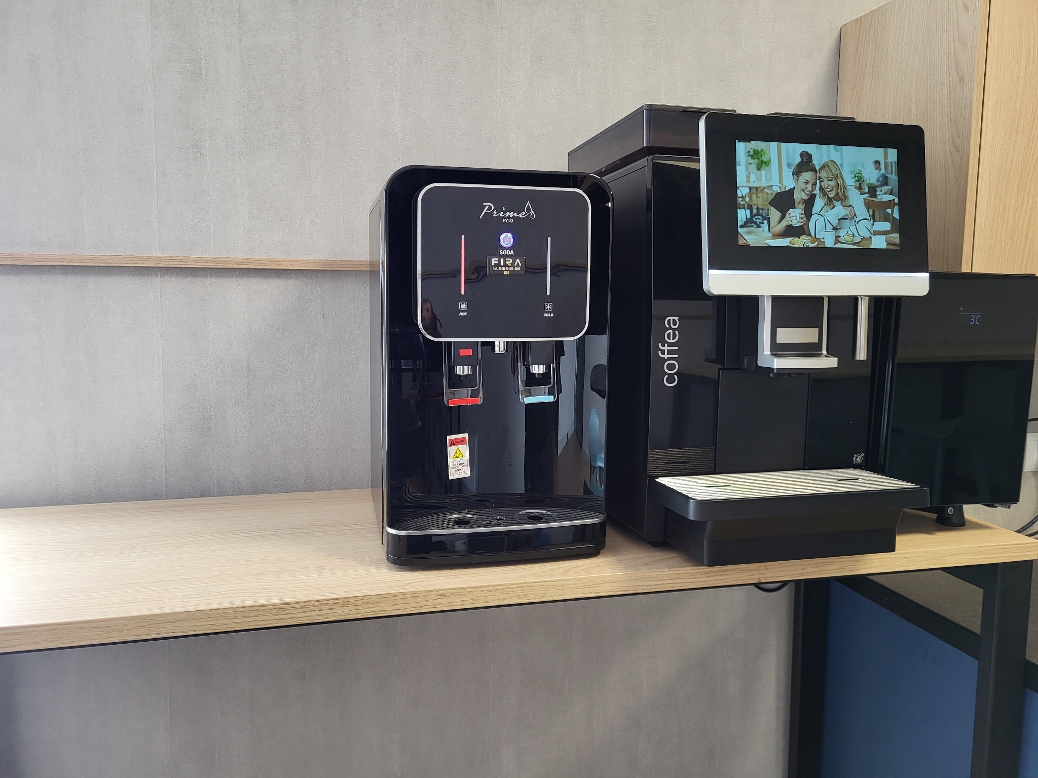 Dwa czarne dystrybutory wody i ekspres do kawy na jasnym blacie, z widocznym logiem 'Prime ECO' na dystrybutorze wody i ekranem z wizerunkiem osób pijących kawę na ekspresie.