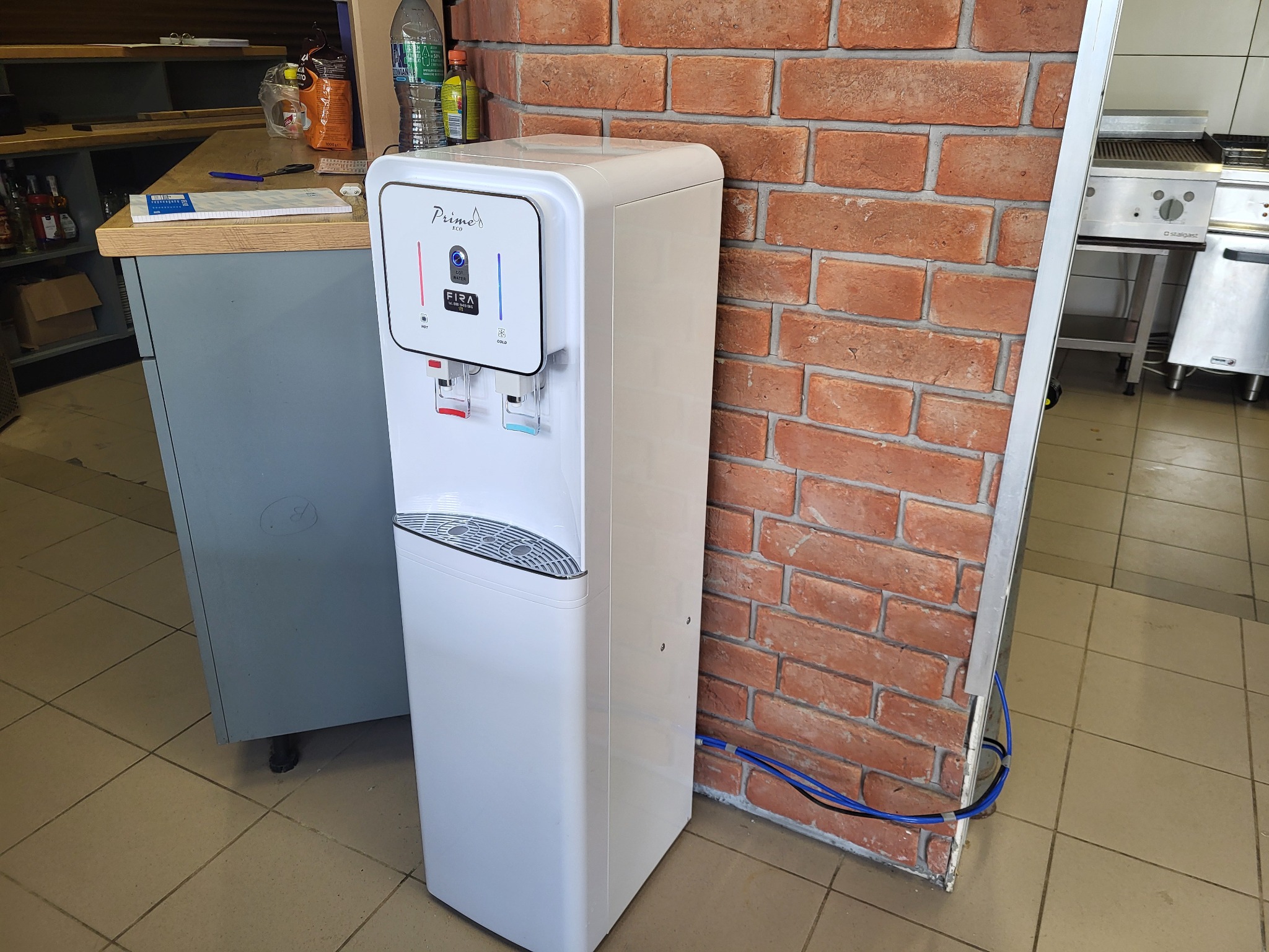 Biały dystrybutor wody marki Prima ECO z podłączeniem do sieci wodociągowej, stojący obok ceglanej ściany i szarej szafki w przestrzeni biurowej lub gastronomicznej.
