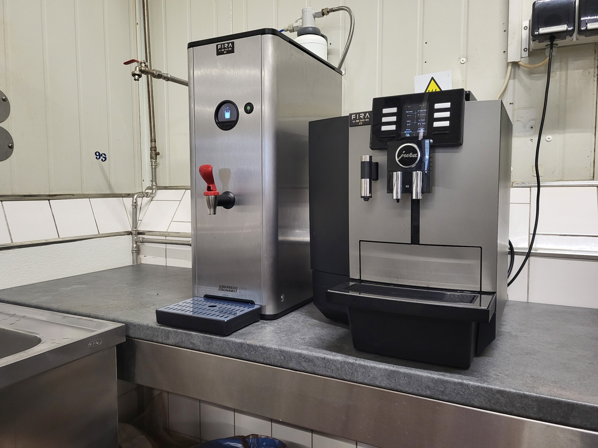 Profesjonalny ekspres do kawy Jura X6 oraz dystrybutor gorącej wody Bravilor Bonamat Fira, ustawione obok siebie na blacie w Słupsku. Dystrybutor wody ma kranik z czerwoną rączką i wyświetlacz...