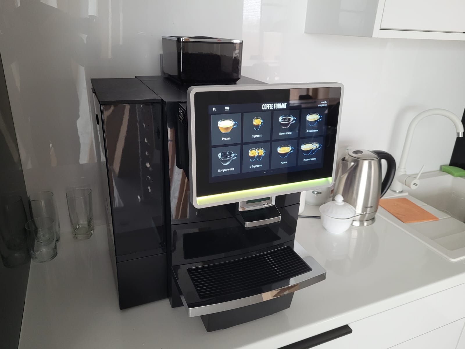 Czarny, automatyczny ekspres do kawy z ekranem dotykowym prezentującym menu napojów kawowych, obok czajnika i szklanek na białym blacie.