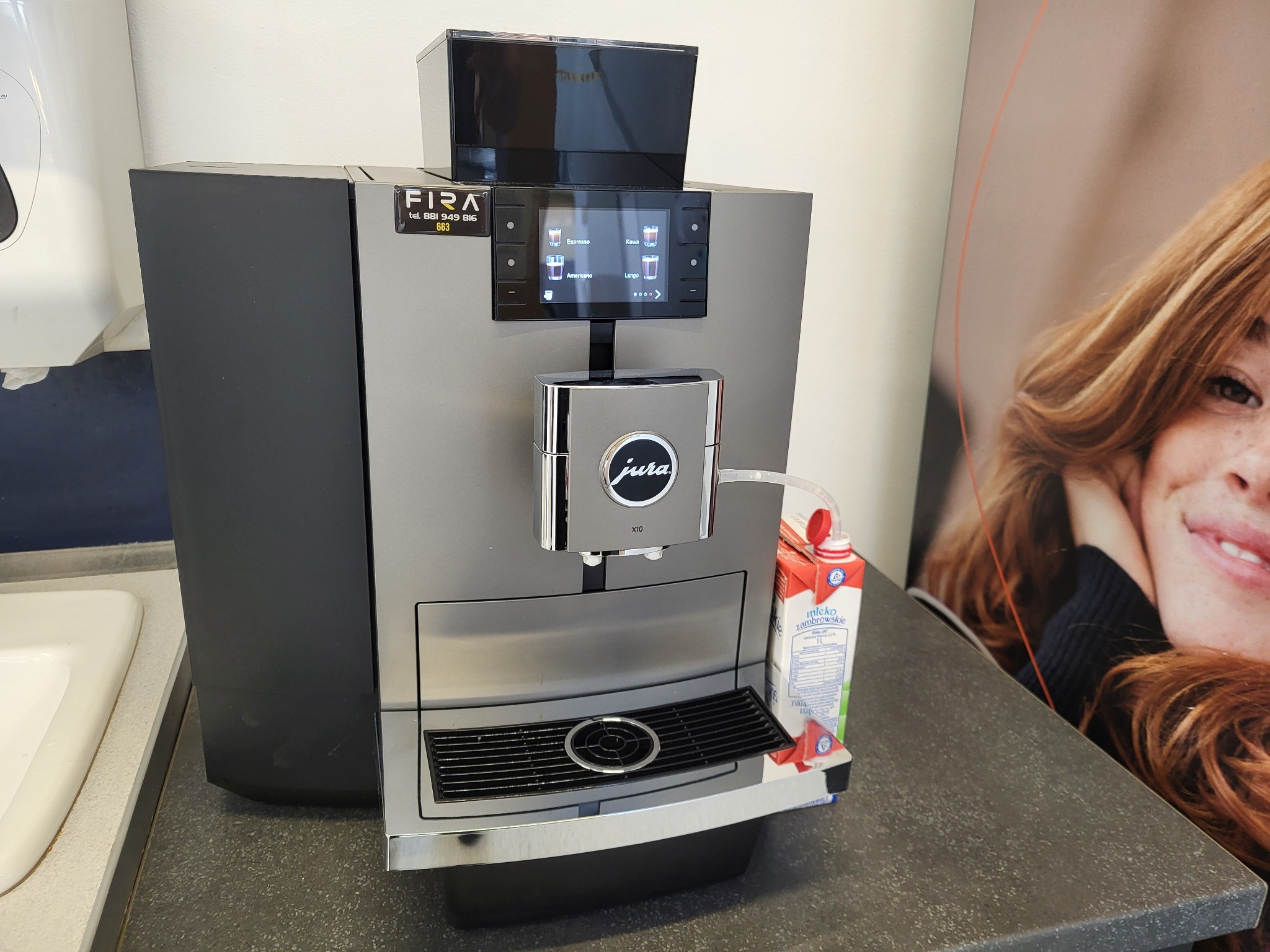 Profesjonalny ekspres do kawy Jura X10 w biurowym otoczeniu, obok karton mleka i plakat z wizerunkiem kobiety.