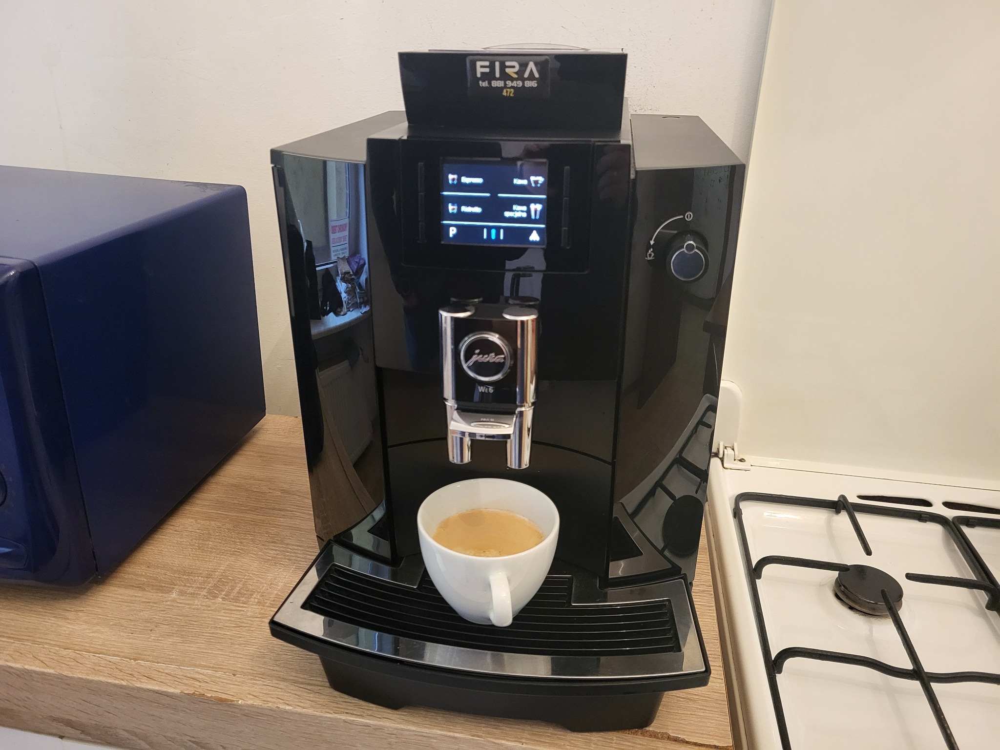 Automatyczny ekspres do kawy Jura W6 z filiżanką kawy na tacce ociekowej, na blacie kuchennym obok kuchenki gazowej i niebieskiej szafki.