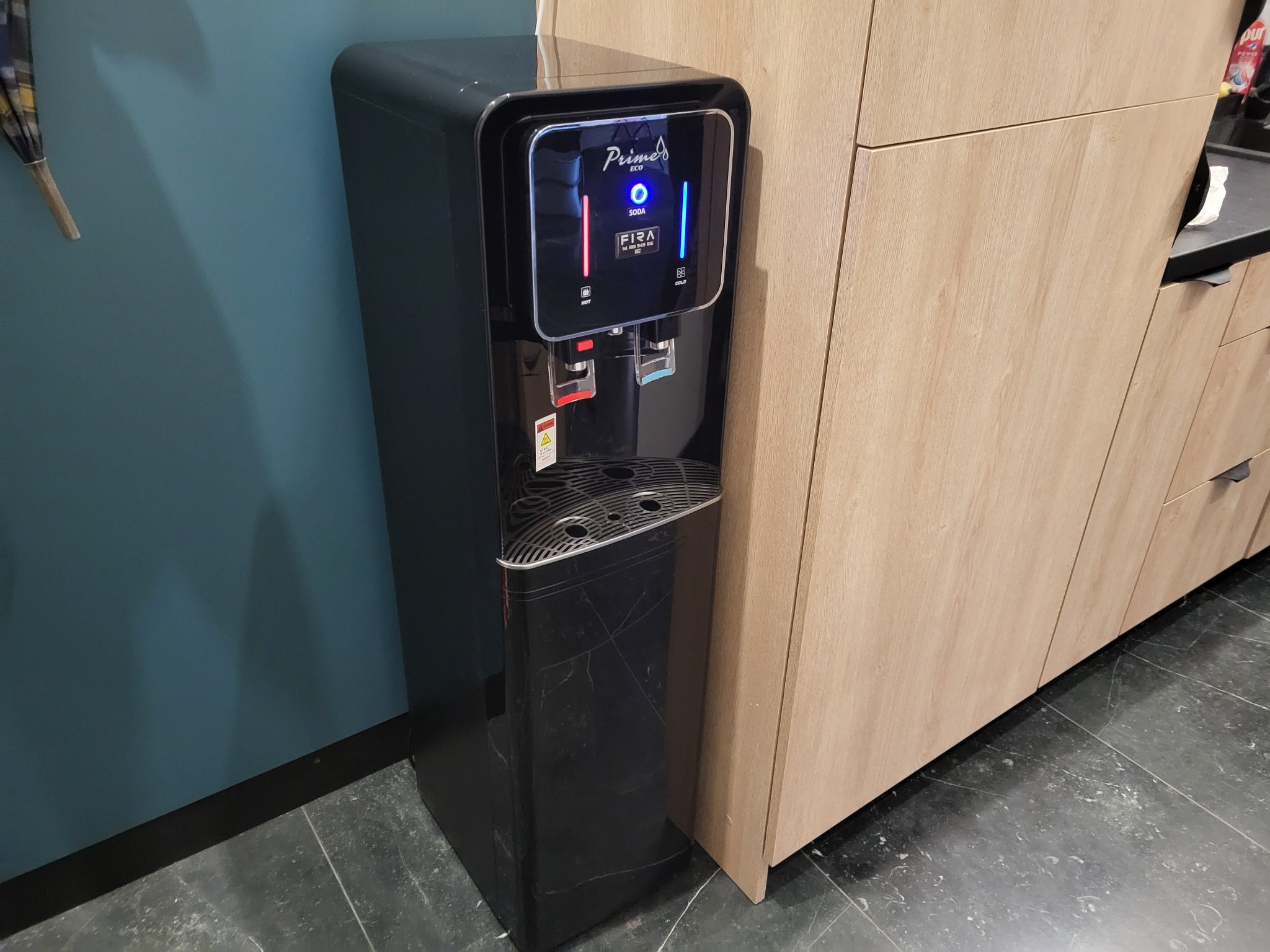 Czarny, wolnostojący dystrybutor wody z wyświetlaczem LED, umieszczony obok drewnianych szafek kuchennych i niebieskiej ściany.