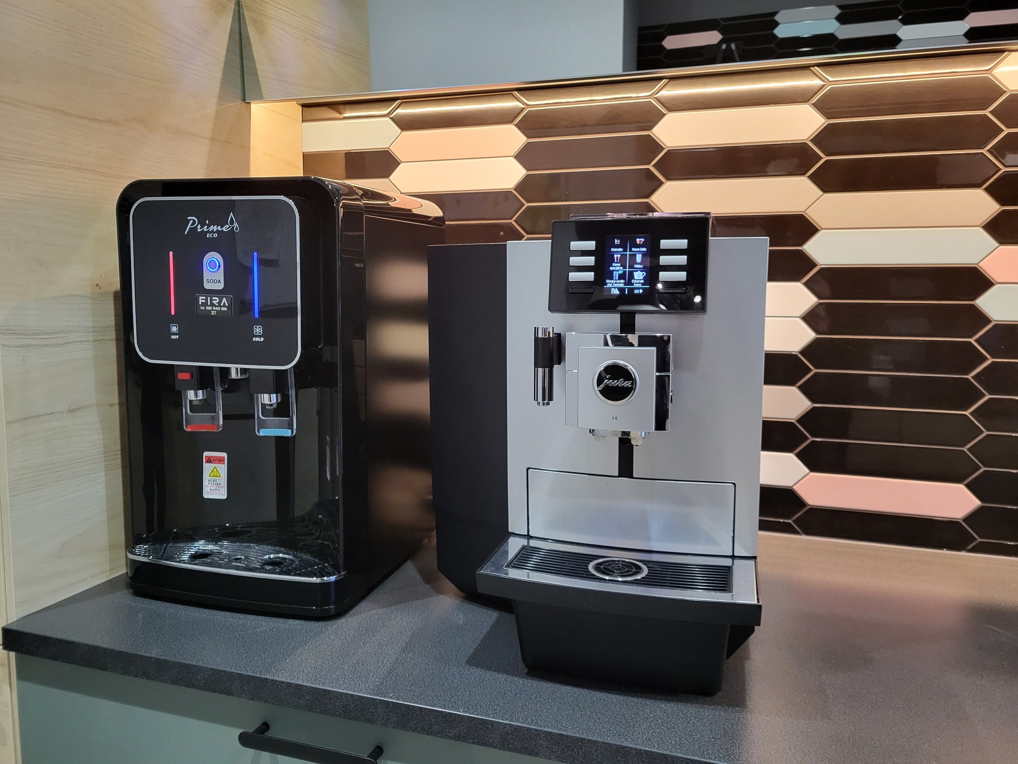 Czarny dystrybutor wody Prime FIRA z podświetleniem LED obok srebrnego ekspresu do kawy Jura XS na blacie z ciemnego kamienia, na tle ściany z dekoracyjnymi płytkami w kształcie rombów w odcieniach...